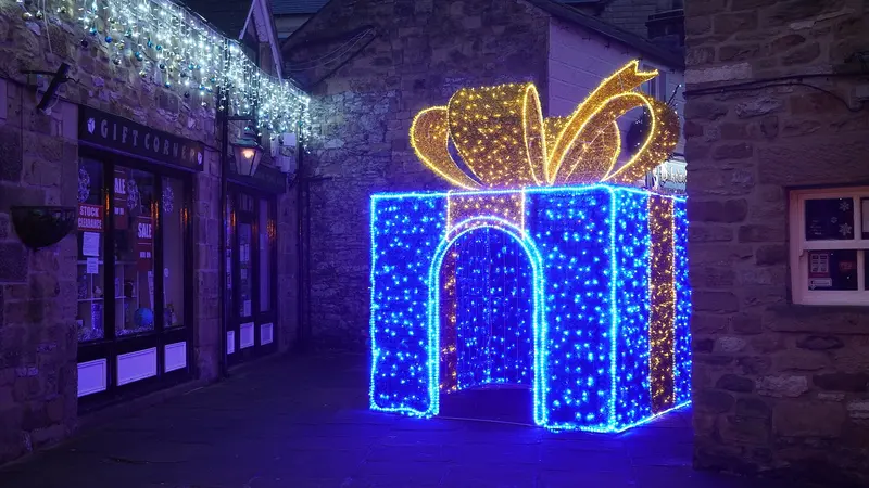 A walk-through light sculpture of a giant blue gift box with a golden bow, beside stone shopfronts hung with icicle lights at dusk