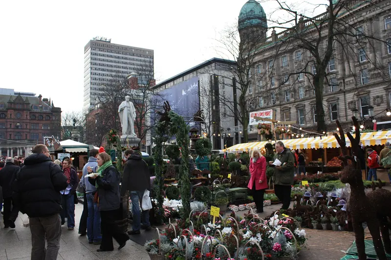 Shoppers walking between Christmas market stalls and a flower stall decorated with a wicker reindeer, with a white statue and the green dome of Belfast City Hall behind