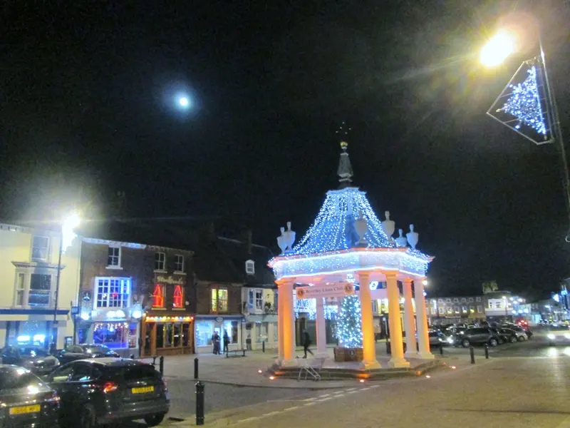 Beverley's market cross outlined in blue Christmas lights at night, with a lit tree beneath it and shops around the square