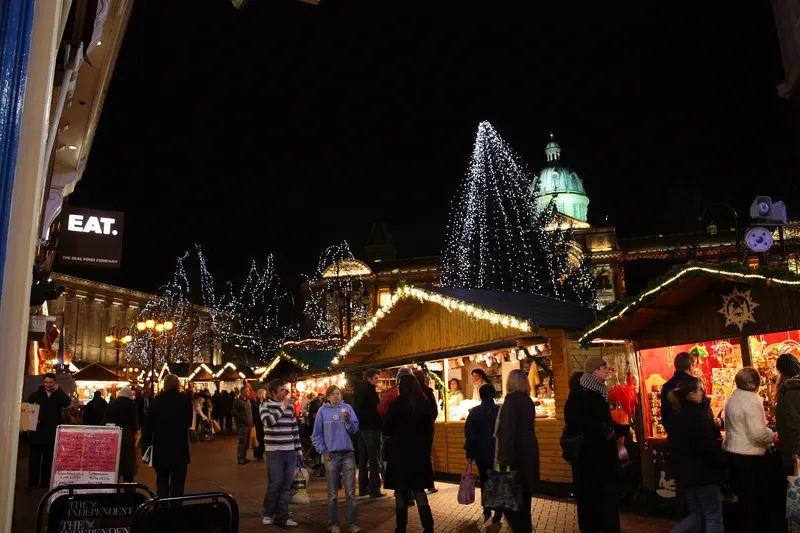 Wooden chalets lit with festive lights in Victoria Square at night, with an illuminated tree and the domed Council House behind