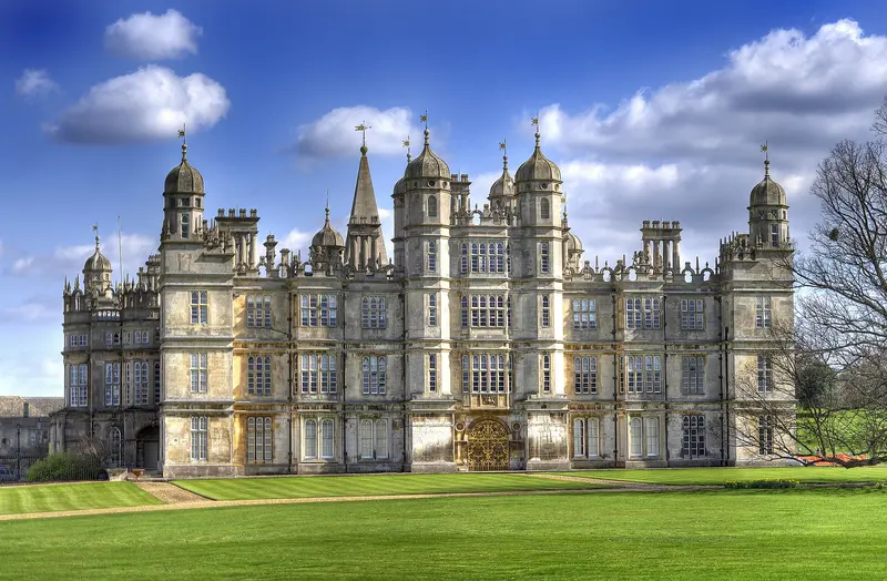 The Elizabethan front of Burghley House, with its pepper-pot turrets and rows of tall windows, above a mown lawn under a blue sky