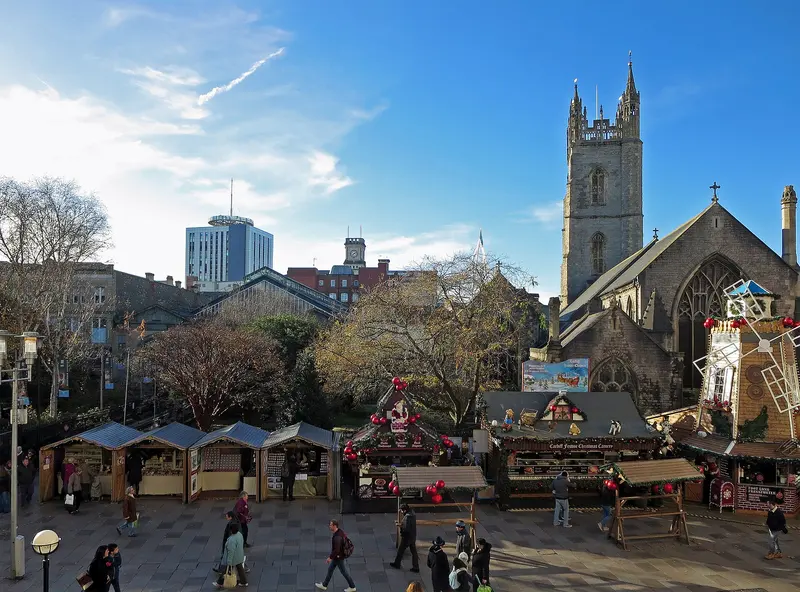 Rows of wooden Christmas market huts and shoppers on Working Street, seen from above, with the tower of St John's Church and autumn trees behind