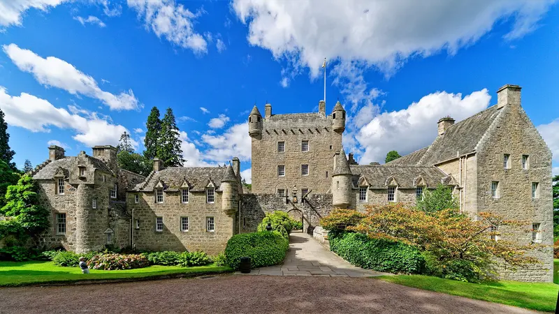 The stone towers and gabled ranges of Cawdor Castle behind flowering shrubs and a lawn under a blue sky with white clouds