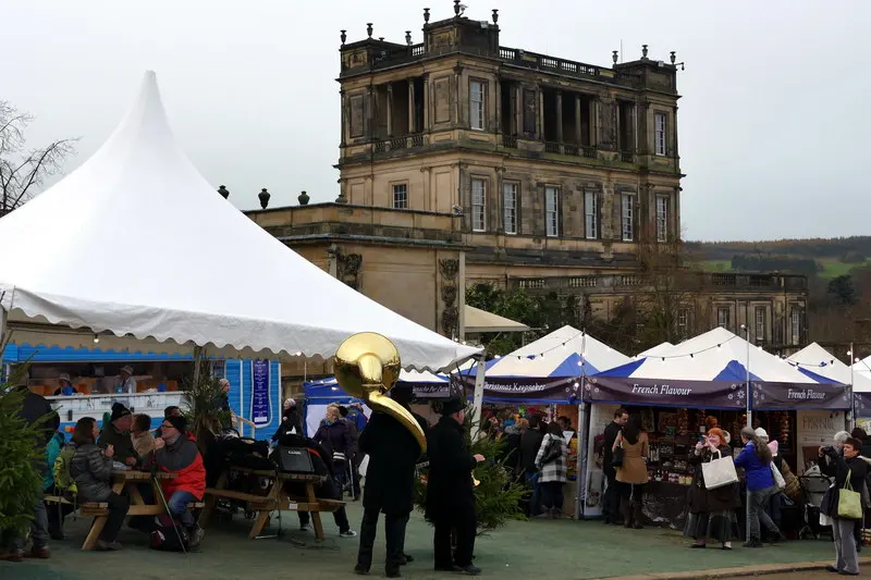 White marquees and blue-and-white market stalls with visitors at picnic tables, below the stone corner of Chatsworth House on a grey day