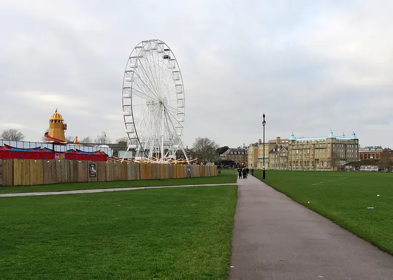 A path across the wide lawn of Parker's Piece towards a big wheel and a helter-skelter behind wooden hoardings, with a large Victorian building to the right
