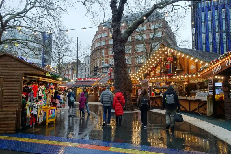 Wooden market chalets strung with warm lights around a large plane tree in Leicester Square, with shoppers on the wet paving