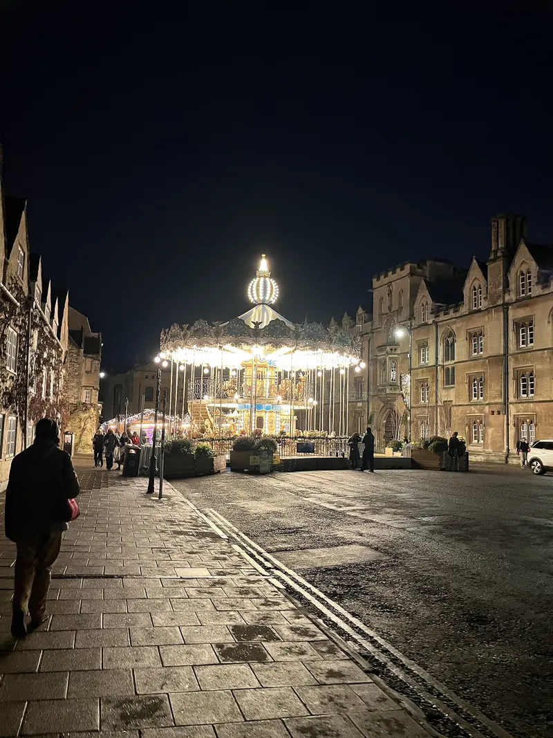 A brightly lit carousel at night at the end of Broad Street in Oxford, between old stone college buildings, with people walking along the wet pavement