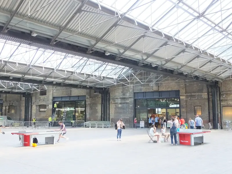 The tall glass-and-iron roof of the West Handyside Canopy at King's Cross over a paved open space, with table-tennis tables and brick buildings beyond
