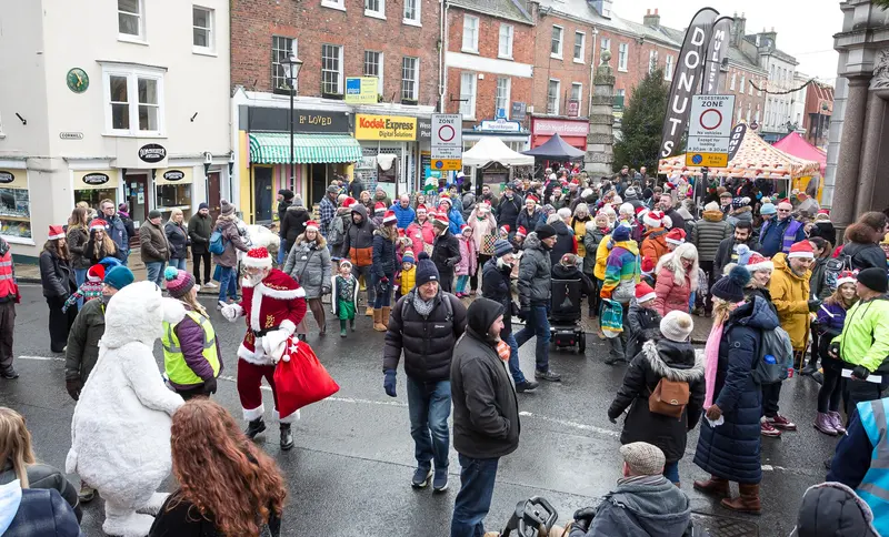 A crowd in Santa hats lines a wet town-centre street as Father Christmas and a polar-bear mascot pass, with gazebo stalls and shopfronts behind