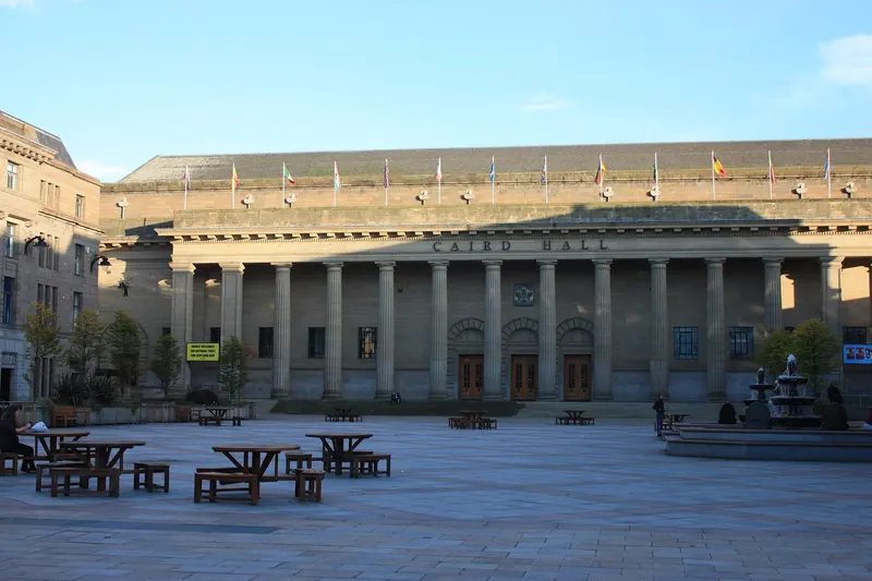 The long colonnaded front of the Caird Hall facing Dundee's paved City Square, with picnic tables and a fountain under a blue sky