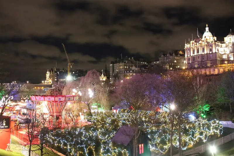 East Princes Street Gardens at night, with fairground rides and market stalls among trees wrapped in coloured lights, below floodlit city buildings