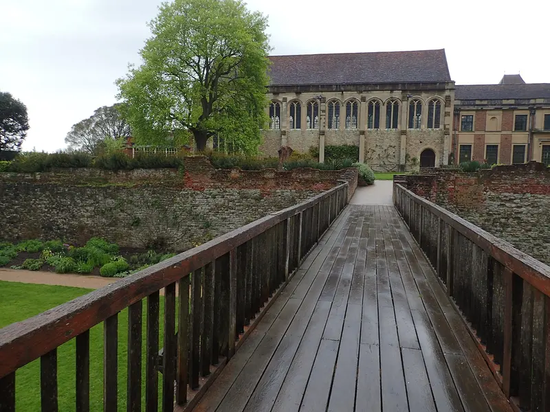 A wooden footbridge across the grassy moat leading towards the medieval Great Hall of Eltham Palace, with its tall windows and steep tiled roof