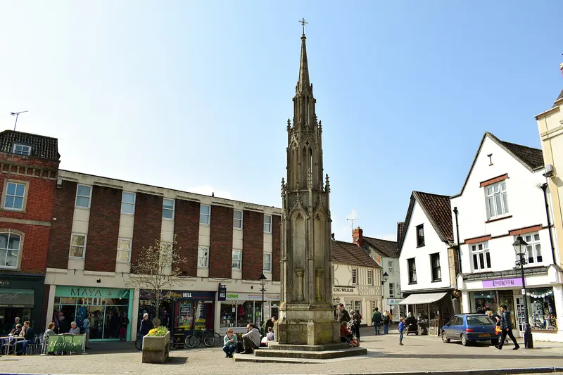 Glastonbury's tall Gothic market cross in the Market Place, surrounded by shops, cafe tables and people out in spring sunshine