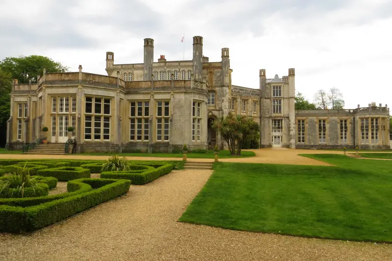 The pale stone Gothic-style Highcliffe Castle with tall pinnacled windows, behind formal box-hedge parterres and lawns under a blue sky