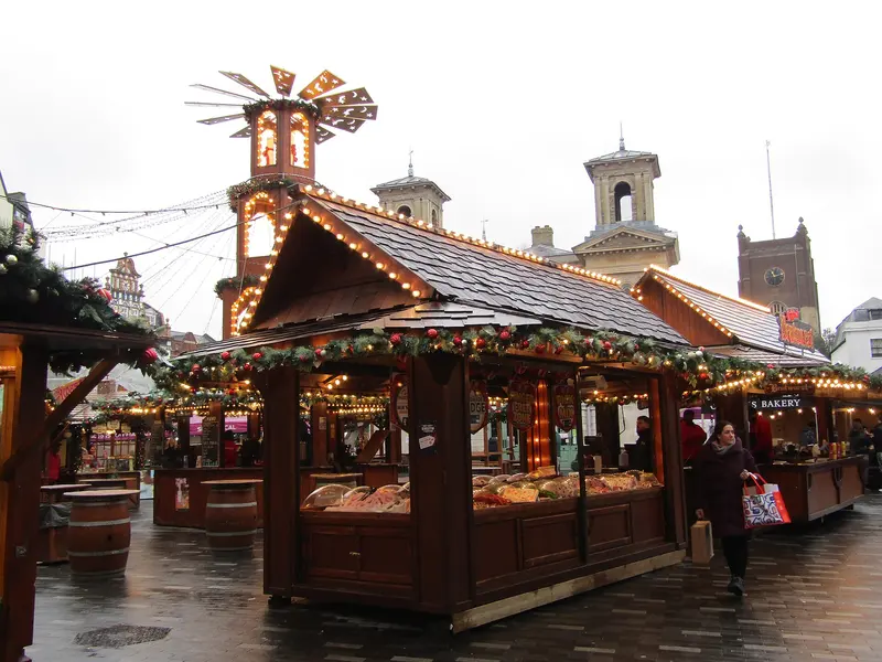 Wooden market chalets trimmed with garlands and lights, with a lit Christmas pyramid and the towers of the old Market House behind