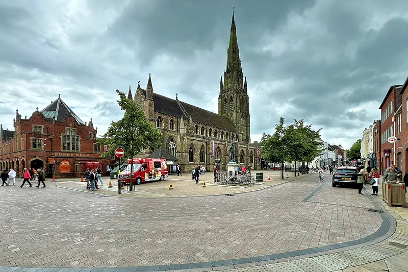 Lichfield's paved Market Square with the spired St Mary's Church, a statue and red-brick buildings around it under a cloudy sky