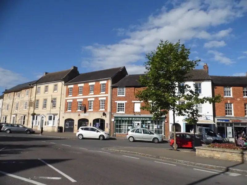 Olney's Market Place lined with Georgian stone and red-brick buildings and a large tree, under a blue sky with white clouds
