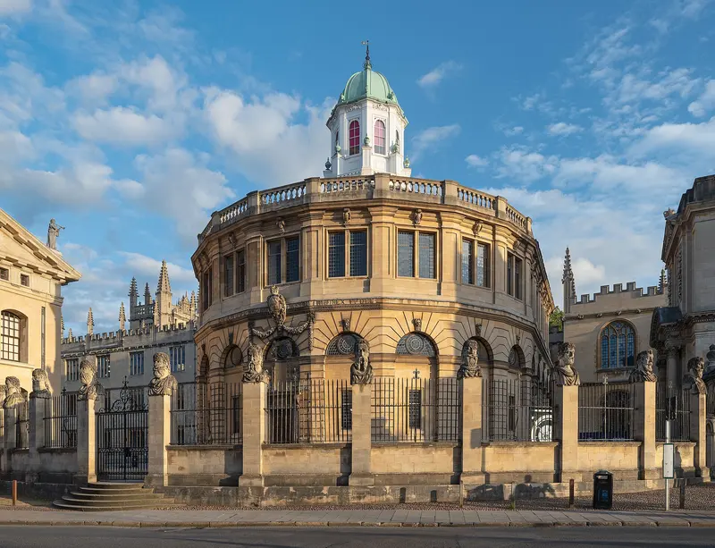 The round, domed Sheldonian Theatre behind railings topped with carved stone heads, on Broad Street under a blue sky