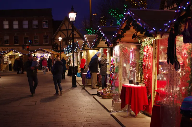 A row of wooden Christmas market chalets trimmed with garlands and coloured lights in Salisbury's Market Place at dusk, with shoppers browsing