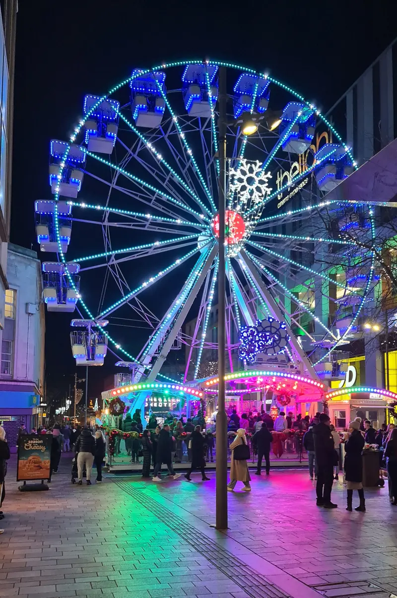 A big wheel lit in blue and pink above a crowd and a brightly lit ride on The Moor in Sheffield at night, with the paving reflecting the colours