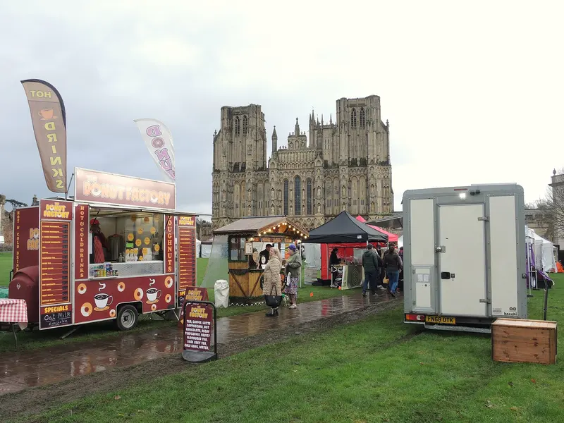Food vans, a wooden chalet and gazebo stalls on the wet grass of Cathedral Green, with the twin-towered west front of Wells Cathedral behind under a grey sky