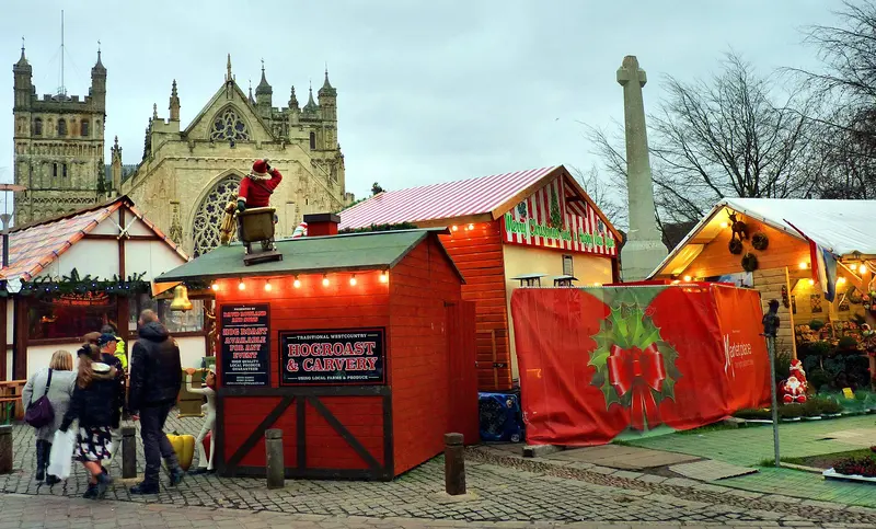 Brightly painted Christmas market chalets with festive decorations and a Santa figure on Cathedral Green, with the towers and west front of Exeter Cathedral behind